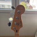 Záběr na hlavu basové kytary z přírodního dřeva s четыřmi chromovanými ladičkami. Na zadní straně hlavy je hnědý nápis Fender a černý text JAZZ BASS®…