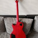Zadní strana červené elektrické kytary s tvarem typu Les Paul. Na zadní straně těla jsou dva černé plastové kryty elektroniky. Kytara má šest ladícíc…