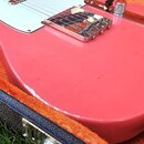 Červená elektrická kytara v oranžovém plišovém kufru. Nástroj má bílý pickguard a chromované kovové části, včetně mostu a knoflíků. Na hraně těla kyt…
