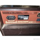 Zadní strana hnědého hudebního zesilovače s červeným zadním panelem. Je zde vidět napájecí konektor AC IN a štítek s nápisy Roland, AC230V~ 50/60Hz 5…