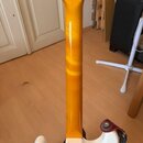 Zadní strana krku a části těla elektrické kytary. Krk je vyroben ze světlého dřeva s žlutým lakem, tělo kytary je bílé. Na spojení krku a těla je vid…