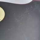Detail tmavého, pravděpodobně černého povrchu hudebního nástroje. V levé části je vidět kulatý, krémově bílý plastový kryt připevněný třemi šrouby. P…