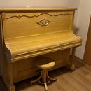 Světle hnědé dřevěné piano s přívodní židličkou. Horní část nástroje je zdobena vyřezávaným ornamentem ve tvaru listu. Povrch dřeva vykazuje známky o…