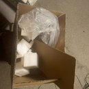 Hnědý cardboardový krabic s vnitřními přepážkami. Uvnitř jsou bílé polystyrenové bloky a průhledný plastový sáček s neznámým obsahem. V horní části f…