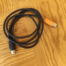 Černý audio kabel svynutý do kroužku, položený na dřevěné podlaze. Na koncích jsou dva různé konektory: jeden černý a druhý oranžový. Na kabelu nejso…