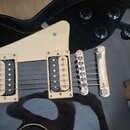 Černá elektrická kytara s bílým plechtrzem a dvěma humbuckery. Nástroj je umístěn v černém kytarovém pouzdře. Na těle jsou vidět čtyři černé ovládací…