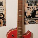 Červená elektrická kytara s tvarem single-cutaway a dvěma humbuckery. Na hlavě je viditelné logo Washburn a grafický symbol kosočtverce. Hриф má hněd…