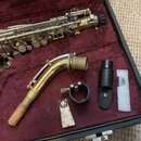 V otevřeném kufru s červeným vnitřkem je zlatý saxofon, jeho hnízdový nástavce a černý mouthpiece s logem KC. K příslušenství patří také černá ligatu…