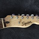 Hlava elektrické kytary z přírodního světlého dřeva s šesti kovovými ladičkami. Na povrchu je černý nápis Squier a pod ním menší text BY FENDER, vpra…