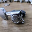 Krytý ladicí mechanismus kytary s chromovaným povrchem. Na boční straně je vyryto logo PRS. V pozadí jsou rozmazané další podobné součástky a malé šr…