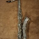 Stříbrný saxofon s černým mouthpiecem. Na těle nástroje nejsou viditelné žádné značky ani nápisy. Povrch vykazuje známky opotřebení a drobné nečistot…