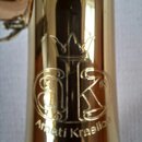 Detail zlatavého kovového povrchu hudebního nástroje. Je zde vyryto logo tvořené korunou nad dvěma propletenými písmeny a nápis Amati Kraslice. Povrc…