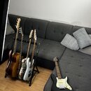 Na fotografii jsou čtyři kytary v černém stojanu a jedna kytara ležící na šedé sedačce. V stojanu jsou zleva hnědá akustická kytara, dvě kytary s pří…