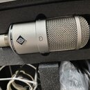 Stříbrný kondenzátorový mikrofon v černé pěnové výplni kufru. Na těle je vidět logo značky a nápis NEUMANN MICROPHONE. V dolní části kufru jsou vidět…