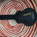 Černý tvrdý kufr na kytaru v akustickém tvaru. Na lidech je zlatým písmem nápis Epiphone. Kufr leží na červeno-bílém koberci s koncentrickými kruhy.