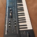 Černý syntezátor s klaviaturaí. Na panelu je viditelný nápis Roland JV-1080 a displej. Nástroj obsahuje množství tlačítek, posuvníků, otočných ovlada…