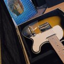 Elektrická kytara s tělem v provedení sunburst a bílým pickguardem, uložená v černém plišovém kufru. Na kytaru je vidět kovový most a ovládací prvky,…