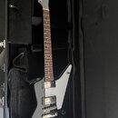 Šedá elektrická kytara s hranatým tělem a bílým pickguardem, položená v černém kufru. Nástroj má dva humbuckery a tři ovládací knoflíky. Na hlavě kyt…