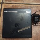 Černý hranatý elektronický přístroj s napájecím adaptérem. V horní části je bílý varovný nápis CAUTION! následovaný textem v angličtině. Povrch příst…