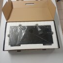 Černý elektronický přístroj v ochranné plastové fólii, umístěný v hnědé kartonové přepravní krabici s bílou vložkou. Na plastové fólii je viditelný r…
