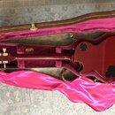 V hnědém kufru s fialovým vnitřkem leží tmavě červená elektrická kytara. Na hlavě nástroje je viditelné logo Gibson. Kytara má černé kruhové krytky n…