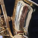 Detail zlatého saxofonu s engravingsm na zvonu. Je zde viditelné logo dvou propletených ladiček a pod ním text YAMAHA ESTABLISHED IN 1887. Povrch nás…
