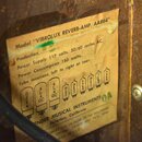 Detail papírové štítku s technickými specifikacemi vylepeného na dřevěný povrch. Na štítku je uveden text: Model "VIBROLUX REVERB-AMP. AA864", Power…
