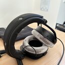 Černé sluchátka s velkými šedými polštářky z plyšového materiálu. Jsou vybaveny spirálovým kabelem a jsou umístěny na dřevěném stole vedle černého te…