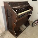 Starý pomalovaný dřevěný harmonium v hnědé barvě. Nástroj má jednu klávesnici s bílými a černými klávesami, nad ní je umístěno sedm bílých ovládacích…