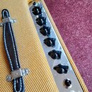 Detail ovládacího panelu zesilovače s kovovým povrchem a žlutým textilem. Panel obsahuje šest černých otočných knoflíků s nápisy VOLUME, TREBLE, BASS…