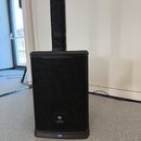 Černý reproduktor s kovovou mřížkou a hranatým tvarem. V dolní části je umístěno logo JBL PROFESSIONAL. Na spodním okraku je vidět malý modrý prvek.…