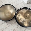Dvě kovové ruční bubny v barvě zlaté s černým okrajem. Pravý nástroj má na povrchu několik vyhlubení v podobě důlků, v horní části je viditelný malý…
