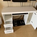Bílý psací stůl s dvěma zásuvkami vlevo a otevřenou skříňkou vpravo, která má rozklopená dvířka. Pod pracovní deskou je umístěna vysuvací police s kl…