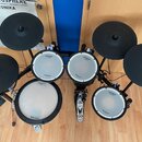 Elektronická bicí souprava značky Roland s řadou padů a činelů V-Drums. Soubor obsahuje čtyři bílé pady s nápisy Roland a V-Drums, tři černé gumové č…