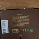 Zadní strana hnědého elektronického zařízení s několika kovovými konektory v horní části. Na povrchu je žlutý text začínající slovem CAUTION a násled…