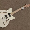 Bílá polodublová elektrická kytara s přírodním hřidlem a hlavou. Na hlavě je nápis "Squier STARSCASTER". Tělo má dva humbuckery, dva ovládací knoflík…
