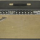 Kytarový zesilovač v černém krytu s béžovou textilní mřížkou. Na ovládacím panelu jsou nápisy Vibrolux Reverb Amp, FENDER MUSICAL INSTRUMENTS a DESIG…