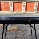 Černý digitální klavír umístěný na černém stojanu. Na zadní straně je bílý nápis SOLTON by KETRON a dále KETRON X1. Na povrchu je viditelná jedna mal…