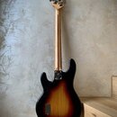 Zadní pohled na elektrickou basovou kytaru s tělem v provedení sunburst (černá, červená a žlutá). Nástroj má světlý dřevěný krk a hlavu s kovovými la…