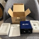 Na šedém křesle je rozložen otevřený hnědý karton s nápisem JET CITY AMPLIFICATION a neúplným slovem Jettenu. Vedle něj je modrý kovový box s bílým l…