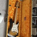 Elektrická kytara s tónem sunburst a bílým pickguardem v otevřeném kufru s oranžovou výplní. Na hlavě kytary je viditelné logo Fender. V kufru je při…