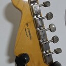 Zadní strana hlavy kytary v přírodním dřevě s lakovaným povrchem. Na zadní straně je černý nápis SERIAL NUMBER JD20003564 MADE IN JAPAN. Na hraně hla…