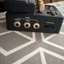 Černý elektronický přístroj s několika zlatými konektory a ovládacími prvky. Na zadní straně jsou tři zlaté vstupy s nápisy Footswitch, Input L (Mono…