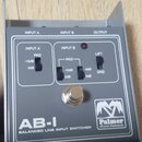 Šedý kovový přístroj s nápisem AB-I BALANCED LINE INPUT SWITCHER a logem Palmer Musical Instruments. Na horní straně jsou označení INPUT A, INPUT B a…