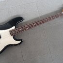 Černá elektrická basová kytara s bílým pickguardem a hnědým hmatníkem. Na hlavě nástroje je viditelné logo Squier a nápis Precision Bass. Povrch kyta…