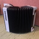 Bílá akordeon s černým měchovcem. Na levém boku je svisle napsáno logo HOHNER a na pravém boku nápis BRAVO. Nástroj vykazuje známky opotřebení v podo…