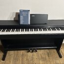Černý digitální piano v černém dřevěném stojanu se třemi pedály. Na klaviatuře leží modrá kniha s nápisy YAMAHA, ARIUS a modely YDP-164, YDP-144 oraz…
