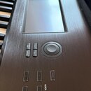 Detailní pohled na ovládací panel digitálního klavíru s kovovým povrchem v kartáčovaném provedení. Panel obsahuje displej, ovládací kolečko a tlačítk…