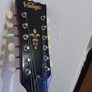 Hlava 12strunné kytary v černé barvě s nápisem Vintage. Uprostřed je umístěn zlatý symbol lilie. Na hlavě jsou viditelné bílé ladičky a struny. Povrc…