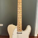 Elektrická kytara typu Telecaster v krémové barvě s bílým plechtrátem. Na hlavě hředu je viditelné logo Fender. Nástroj má javorový hmatník a dvě sní…